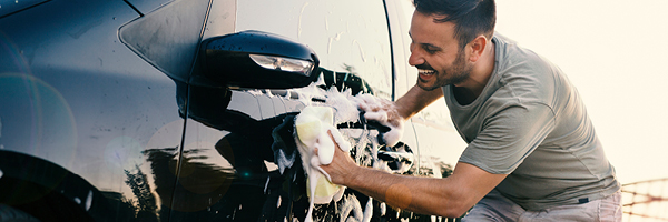 A man in shorts and a t shirt smiles while he washes the side of his black vehicle with a soapy rag.